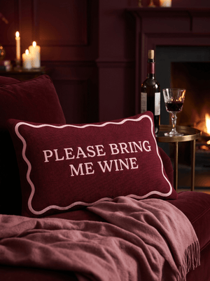 Please Bring Me Wine needlepoint cushion on a burgundy velvet sofa by candlelight